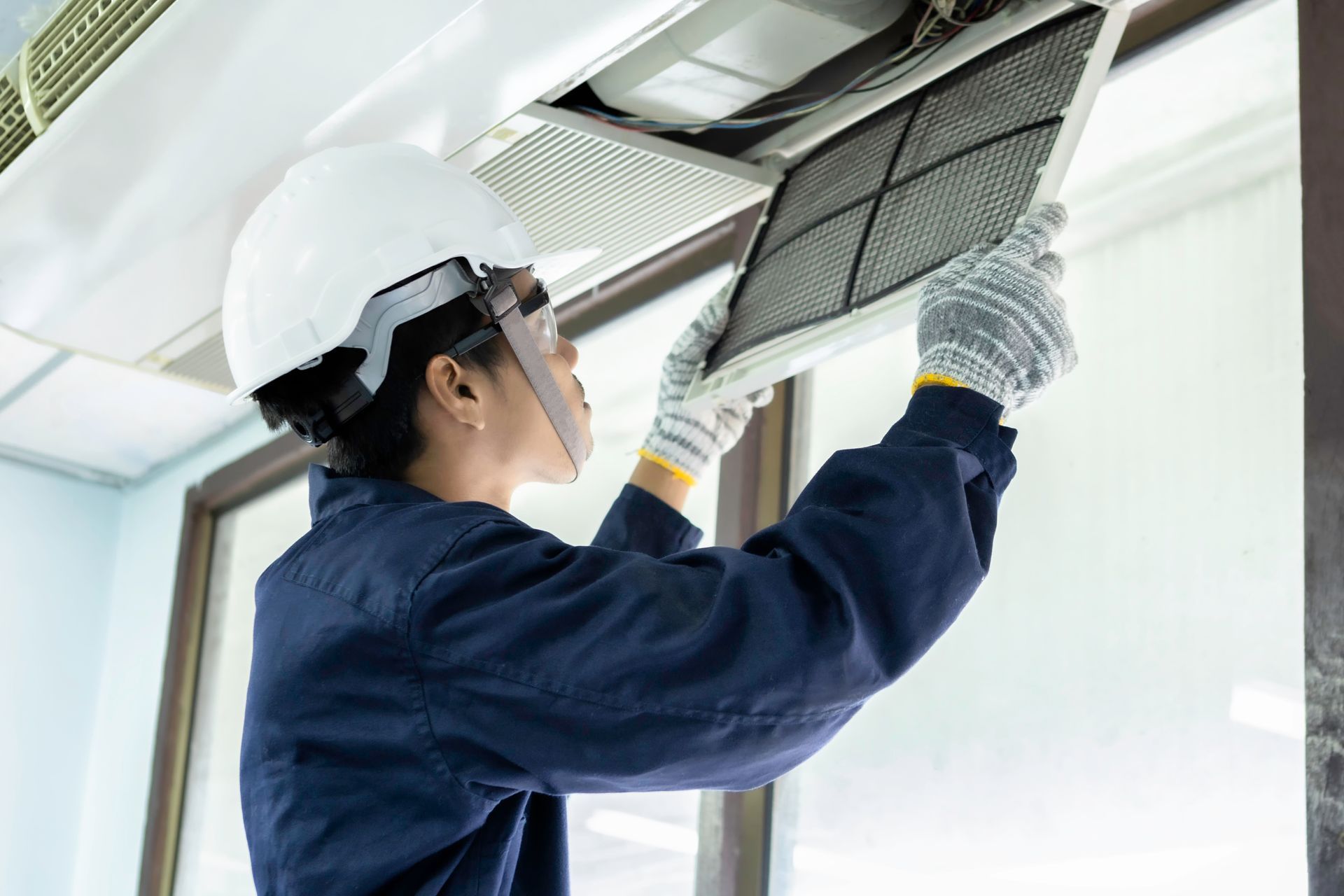 A worker in a white hard hat and blue uniform inspects or replaces a ceiling-mounted air conditioning filter.