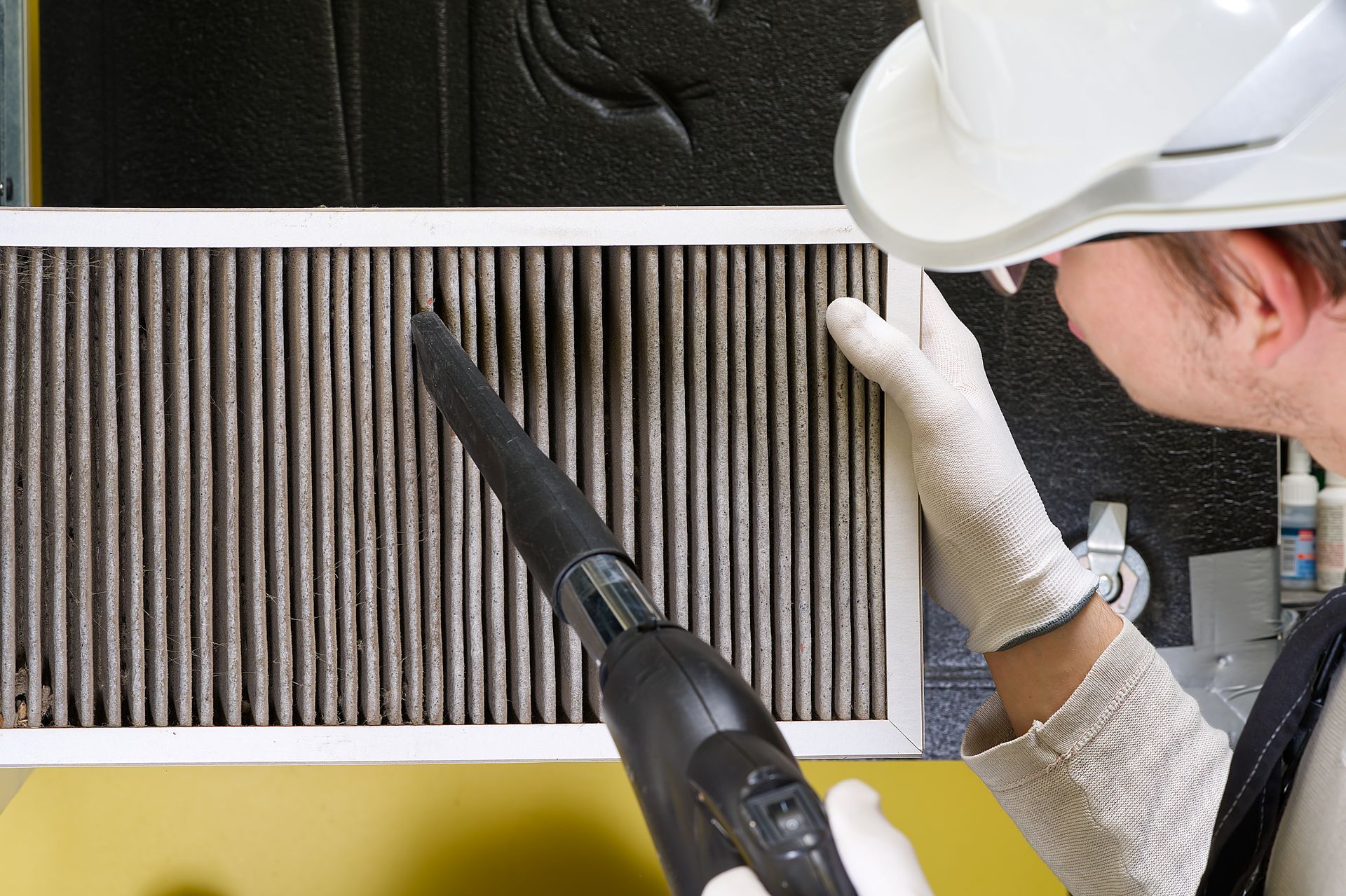 A person wearing a white hard hat and gloves uses a vacuum nozzle to clean a dirty, pleated HVAC air filter.