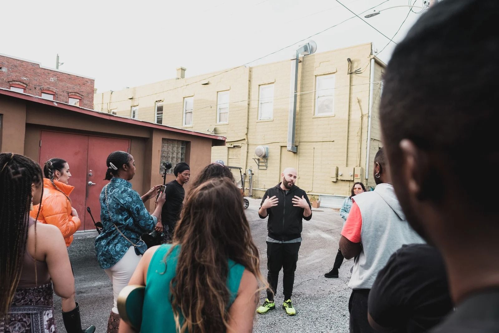 A person gesturing while speaking to a group in an outdoor alleyway near a beige building.