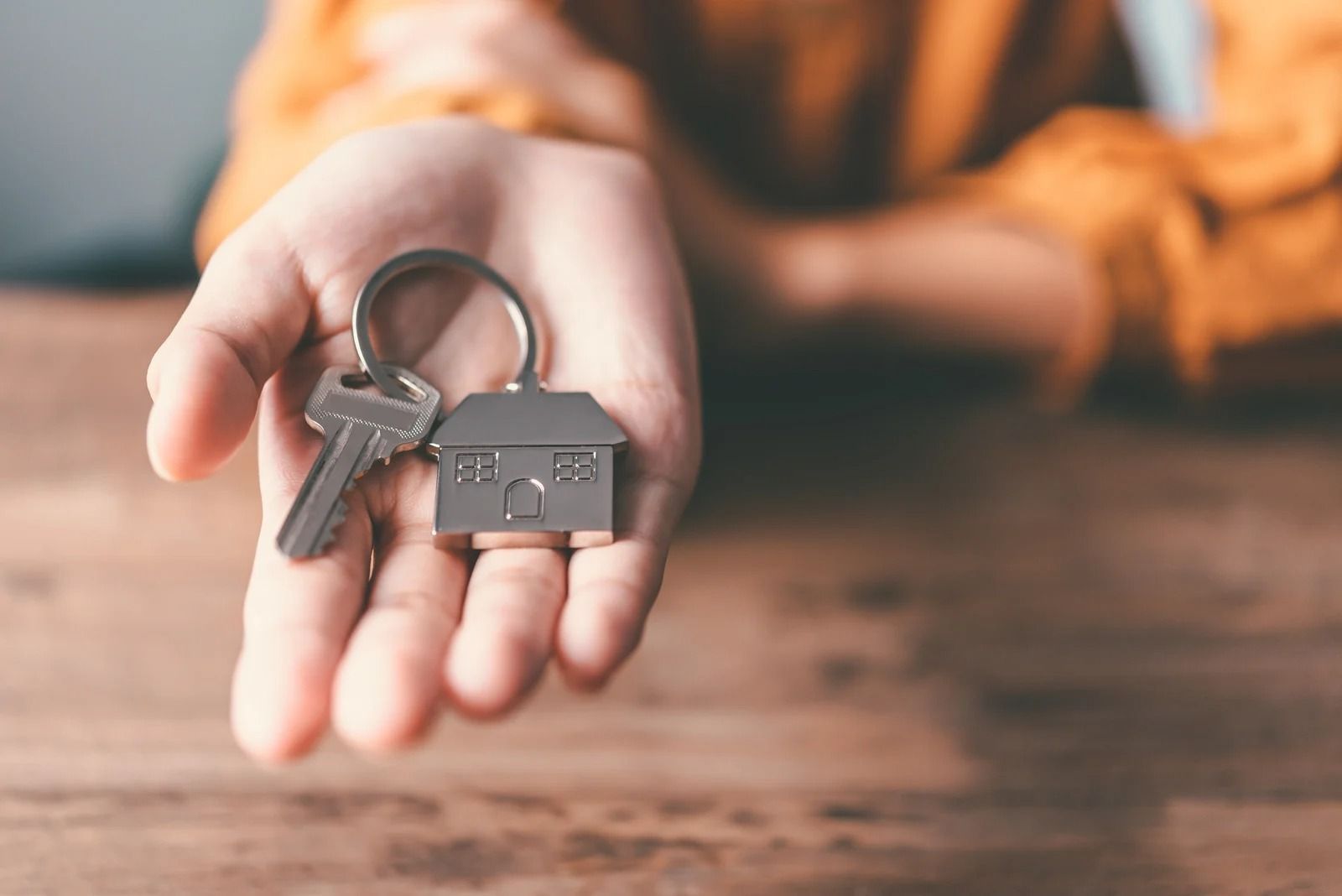 A hand holds a silver key on a keychain shaped like a house, resting on a wooden surface.