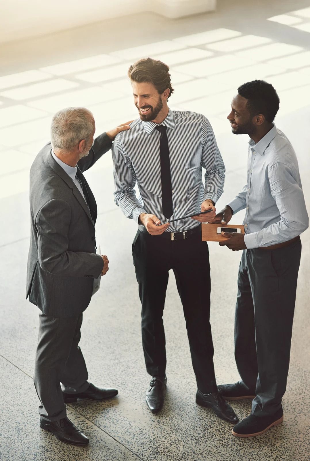 Three professionals in business attire stand in a sunlit office, smiling and conversing while one gestures to another.