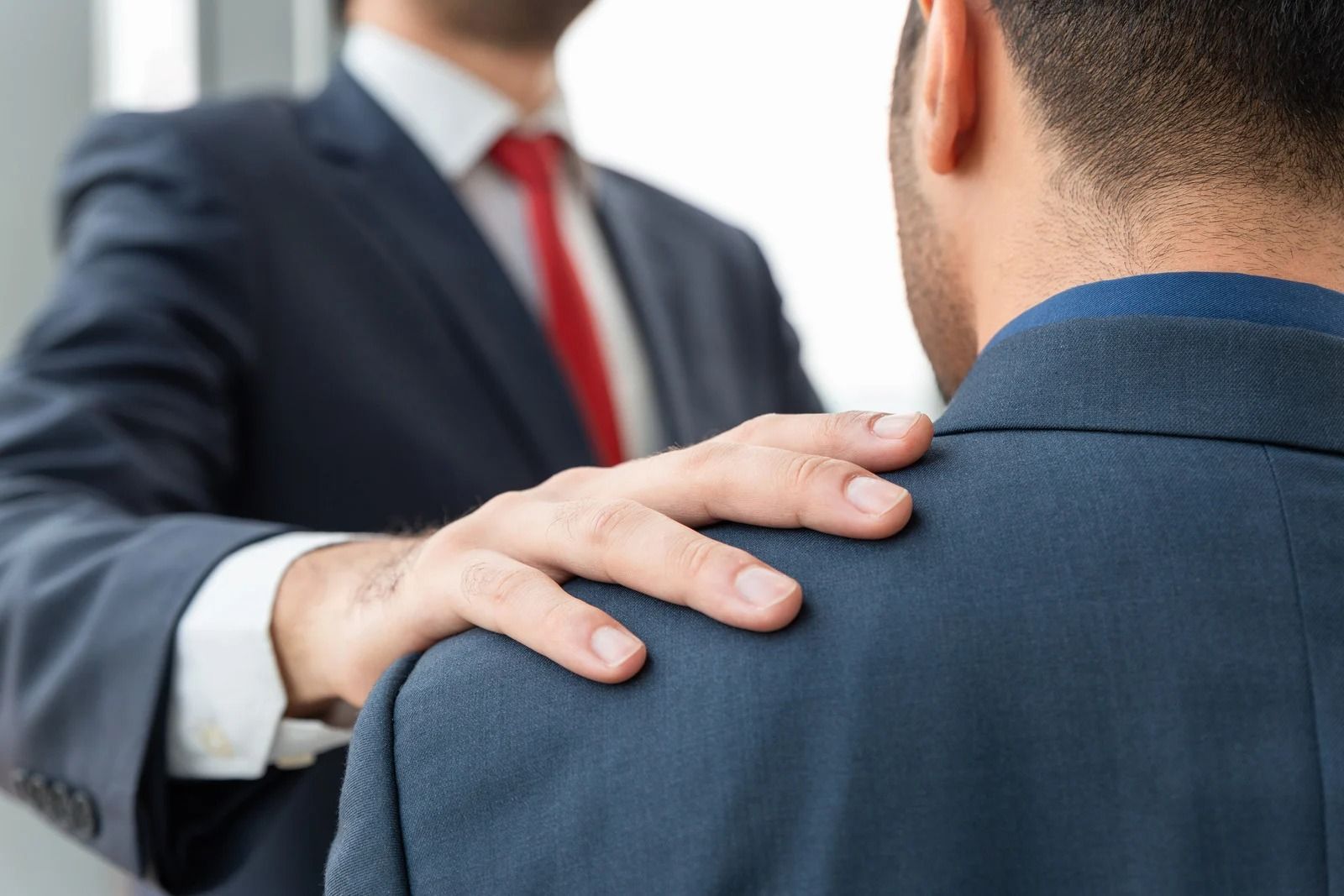 A person in a business suit rests a hand on the shoulder of another individual wearing a dark blazer.