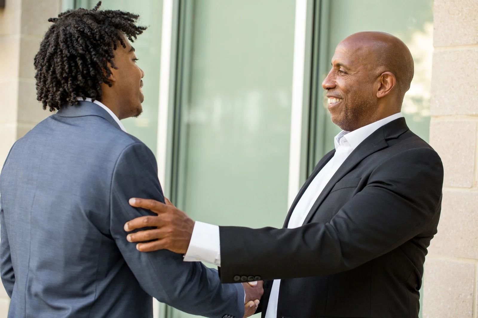 Two business professionals in formal suits shaking hands and smiling while standing outside.