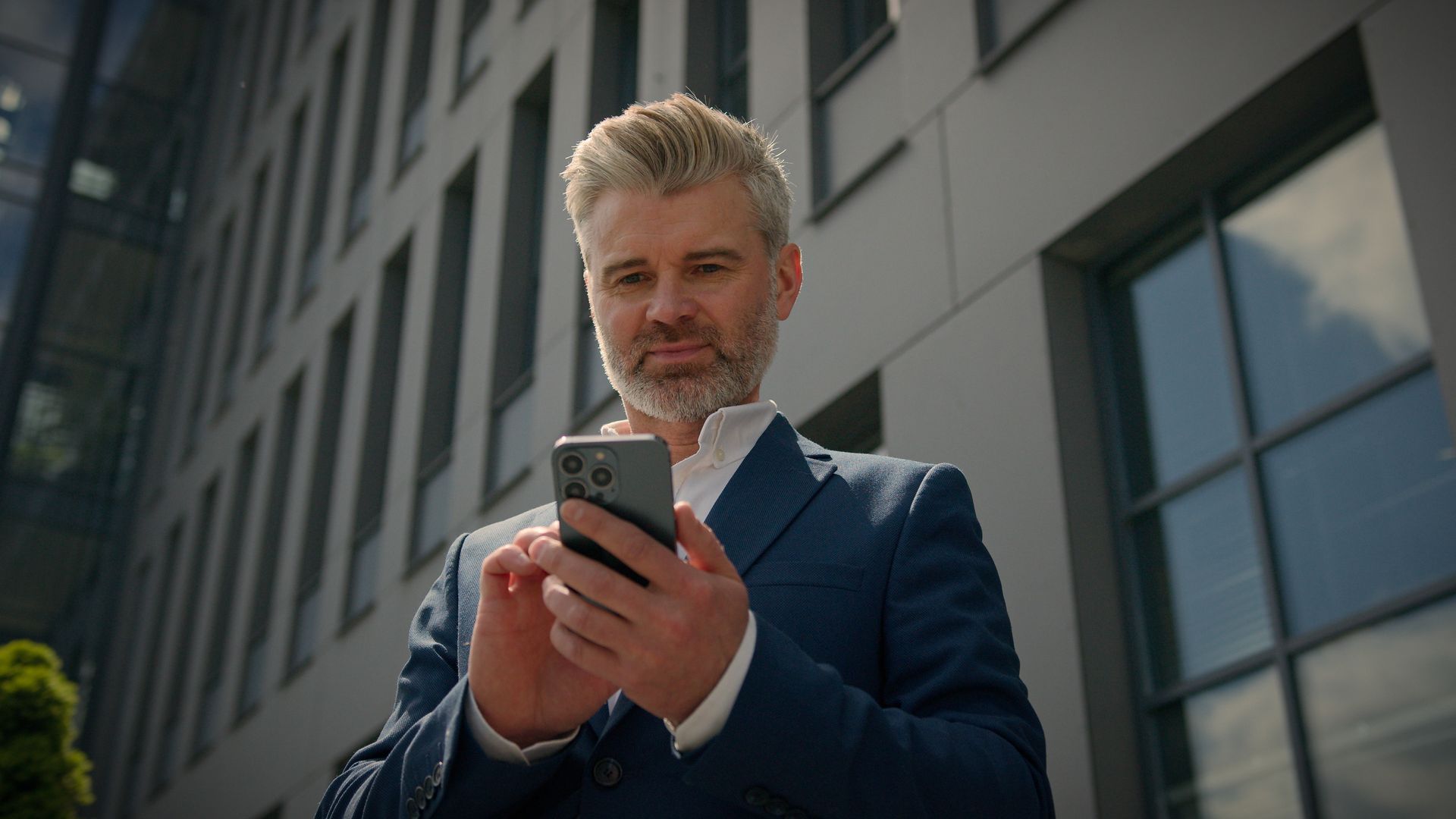 Man in blue suit using a smartphone outside a modern building.