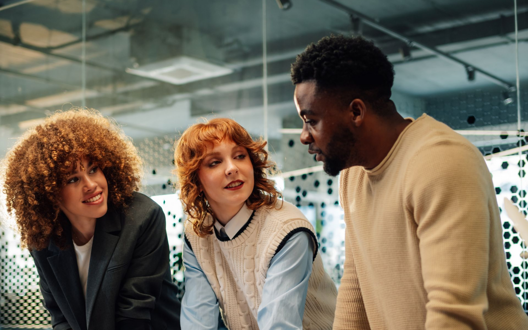 Three people in an office setting. Two women and a man are conversing by a glass wall.