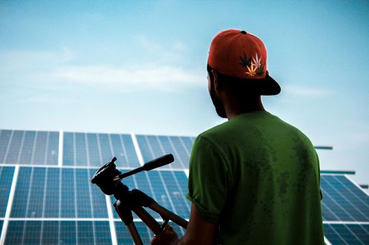 A person wearing a cap and green shirt holds a tripod, looking at solar panels.