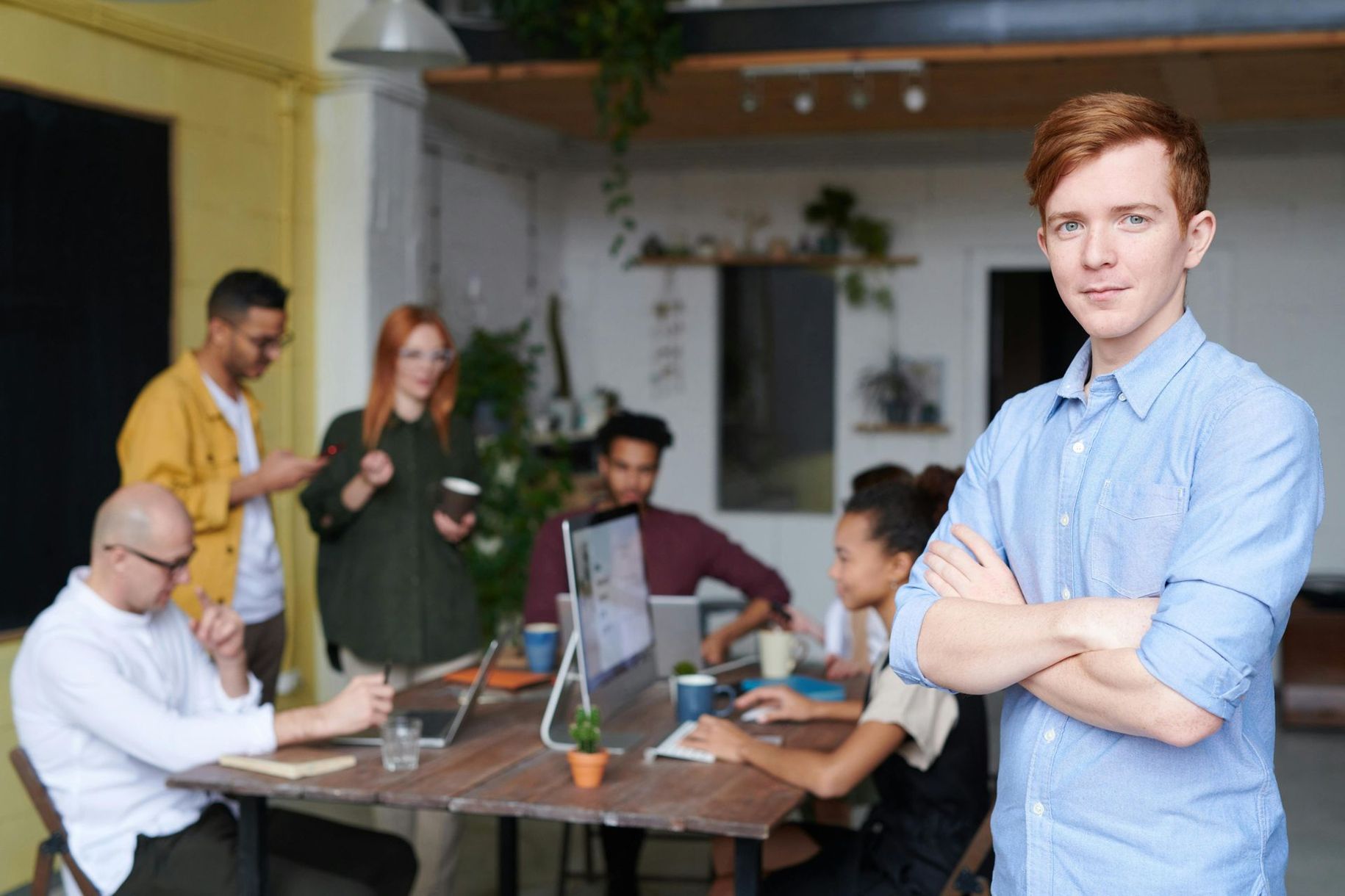 Man in blue shirt with arms crossed, standing in an office with a team working at a table.