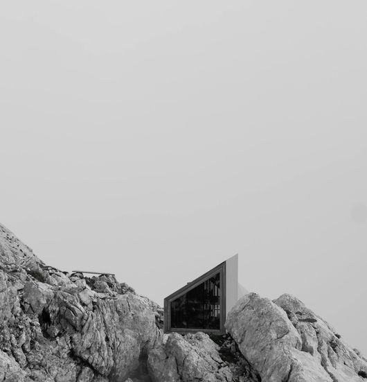 A modern, triangular mountain hut with a large window perched on a rocky mountain peak. The scene is monochromatic.