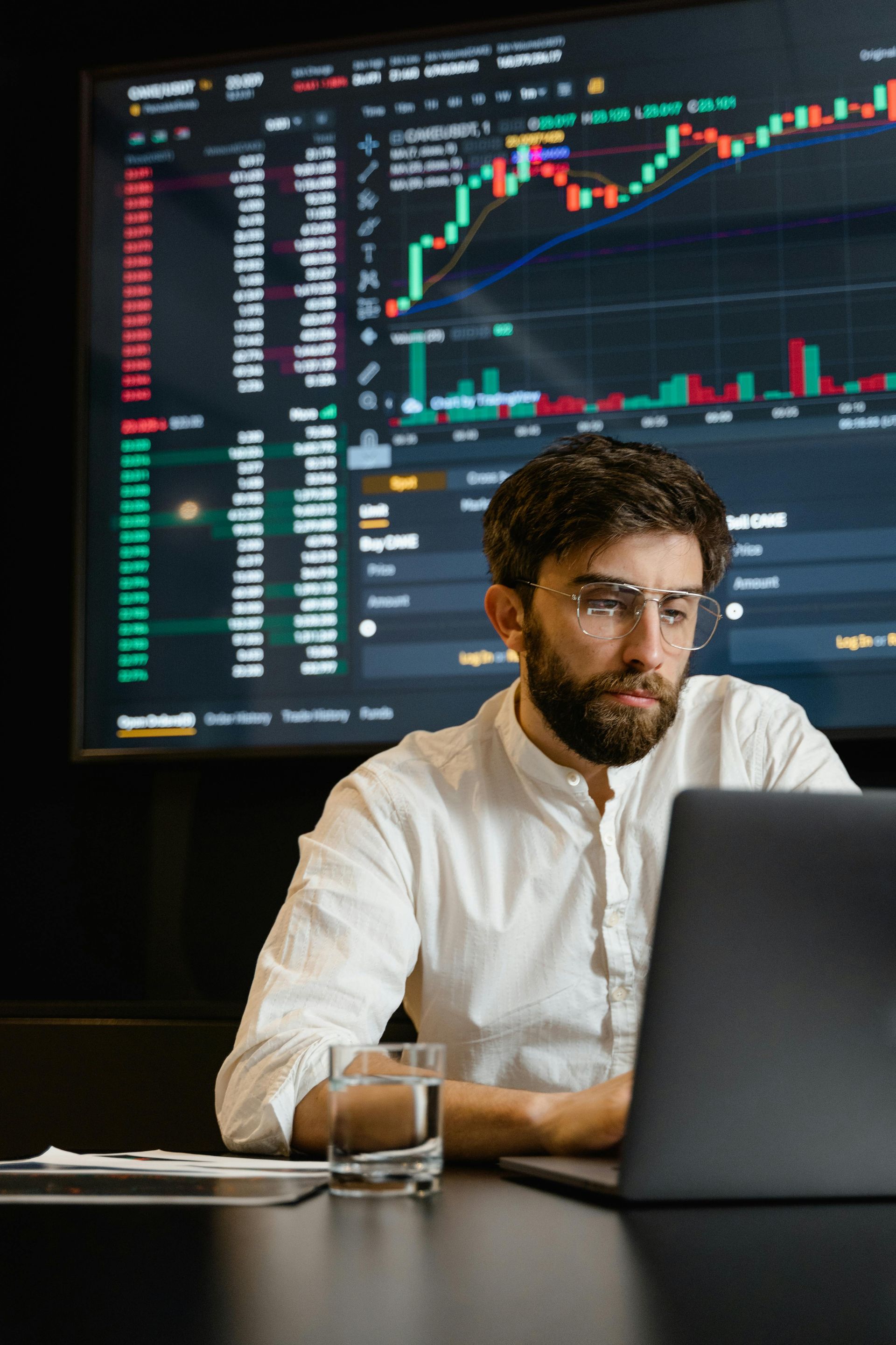 Man using laptop, stock charts on a screen in the background. He wears glasses, a white shirt, and has a beard.