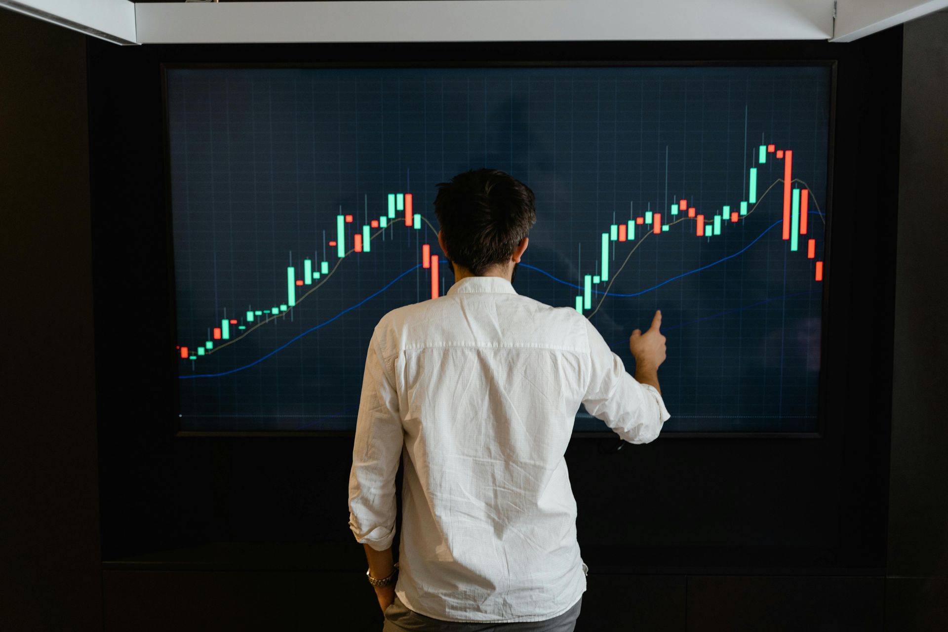 Person points at a stock chart with green and red lines on a large screen.