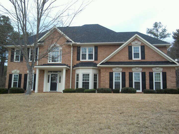 Two-story brick house with black shutters and a dark roof on a grassy lot. The sky is overcast.