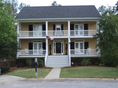 Two-story yellow house with white balconies, black shutters, and a black door; a sidewalk leads to the front door.