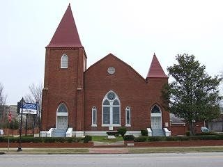 Red brick church building with two towers and red conical roofs. Two white doors with arched tops are in the facade.