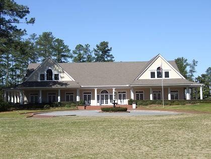 A large, light yellow building with a long porch, set on a grassy lawn with trees in the background under a clear blue sky.