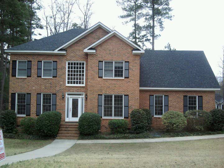 Two-story brick house with black shutters and roof, a white front door, and green bushes in front.