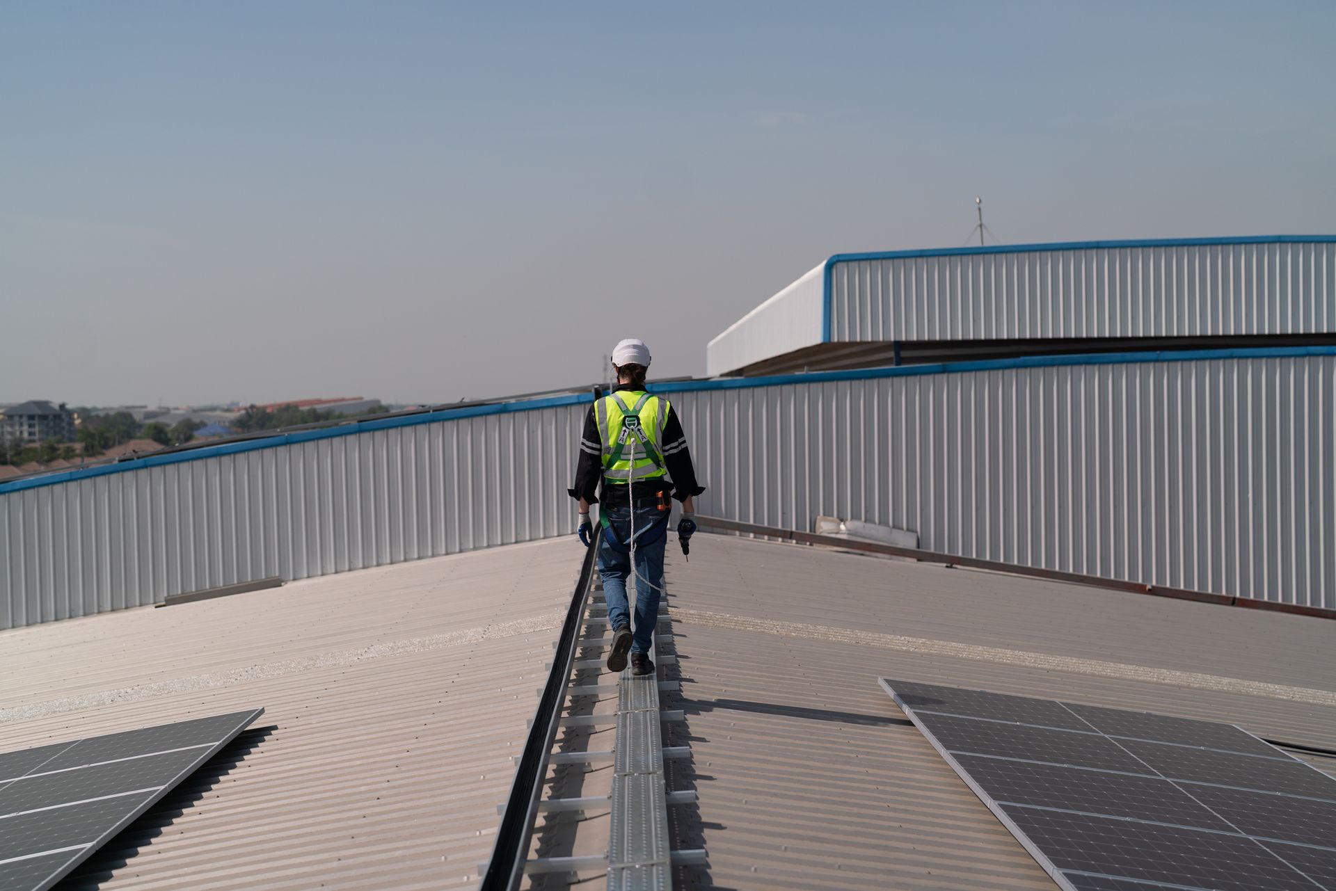 Commercial roofer walking on metal roof with safety harness and solar panels in view.