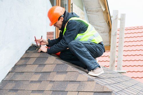Roof Installations — Worker Installing New Roof Shingles in Augusta, GA Roofer in orange hardhat and safety vest repairs shingles on a pitched roof, using pliers.