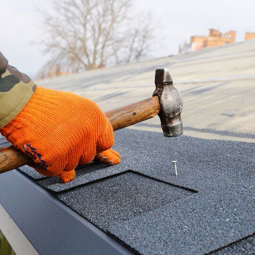 Hand in orange glove hammers a nail into a dark grey asphalt shingle on a roof.