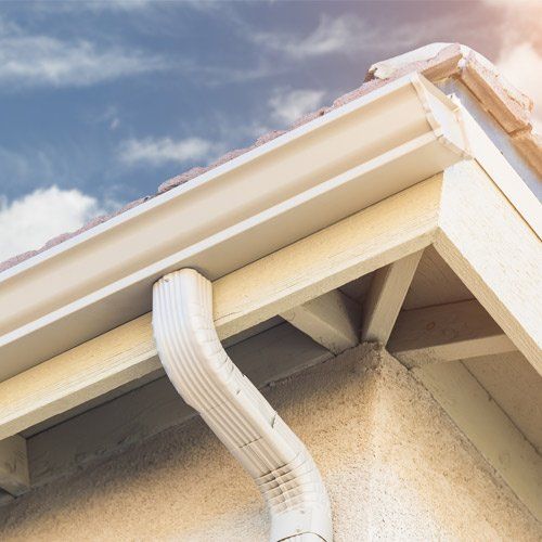 Beige gutters and downspout on a light-colored house, against a backdrop of a partly cloudy blue sky.