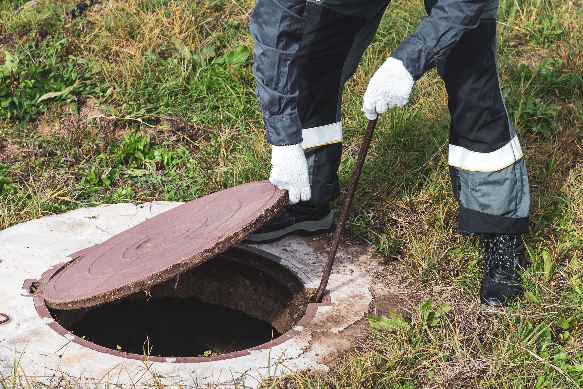 Person in dark work clothes opens a manhole cover with a hook in a grassy area.