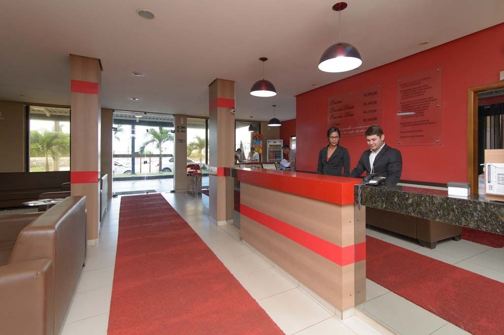 Balcão de recepção do hotel com dois funcionários. Esquema de cores vermelho e marrom com tapete vermelho.
