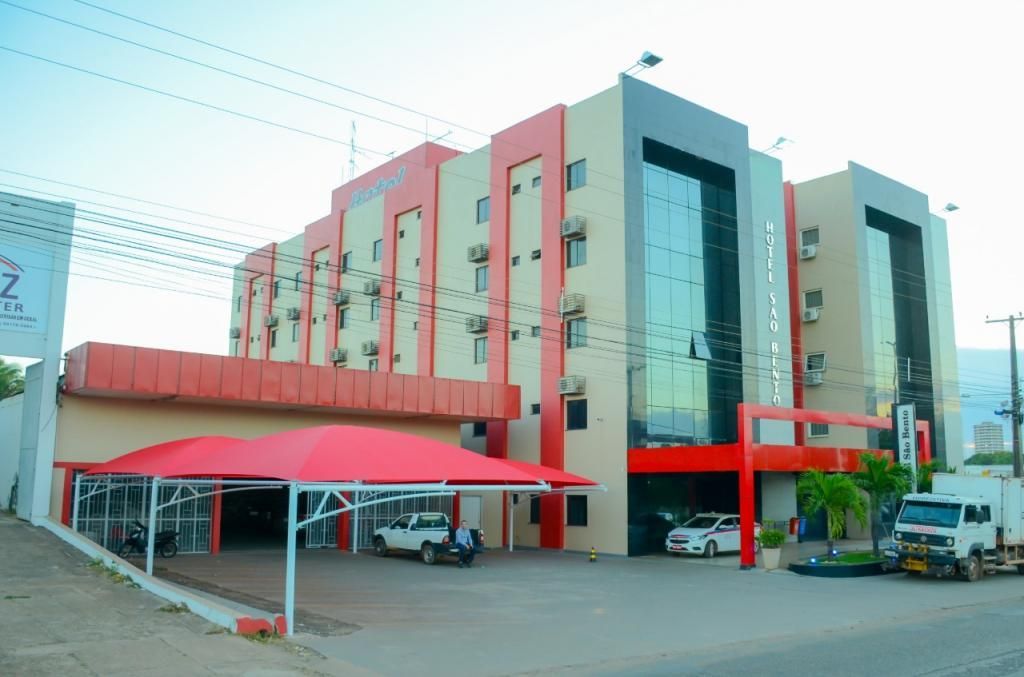 Prédio de hotel com fachada vermelha e bege, frente de vidro e veículos estacionados em frente.