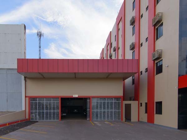 Entrada do prédio em tons de bege e vermelho, com portas de garagem abertas e vagas de estacionamento.