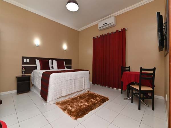 Quarto com cama de casal, cortinas vermelhas, uma pequena mesa com cadeiras e um tapete marrom.