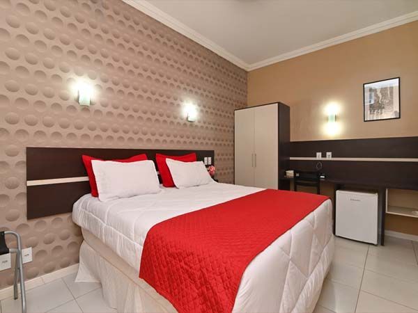 Quarto de hotel com cama queen-size, detalhes em vermelho e frigobar.