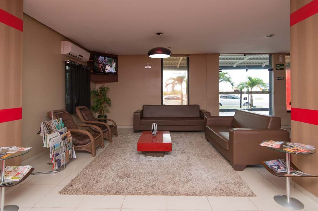 Sala de espera com sofás marrons, cadeiras de vime, uma mesa de centro vermelha e uma TV; revistas em mesas laterais.