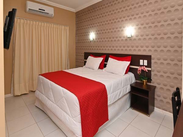 Quarto de hotel com cama vermelha e branca, cortinas creme e papel de parede estampado.