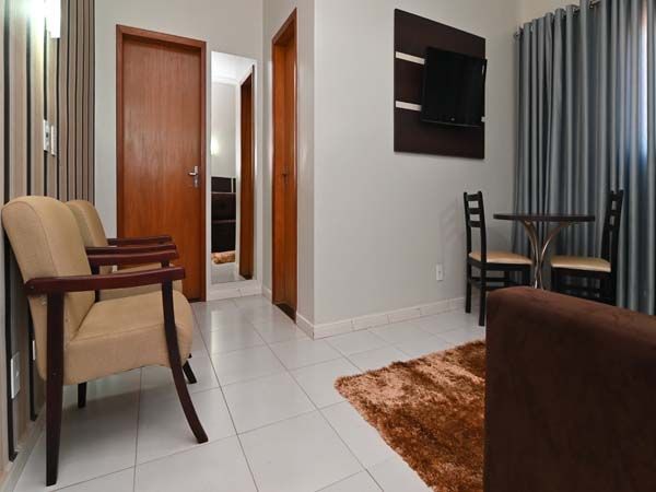 Sala de estar com sofás bege, mesa, TV e portas abertas para outros cômodos. Tapete marrom e piso de azulejo branco.