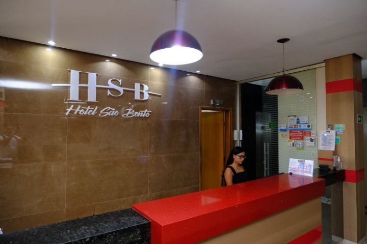 Hall de entrada do hotel com balcão de recepção e uma mulher atrás dele. Placa 