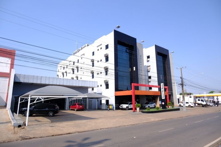 Prédio moderno de vários andares, branco com detalhes em preto e estacionamento, localizado em uma rua ensolarada.