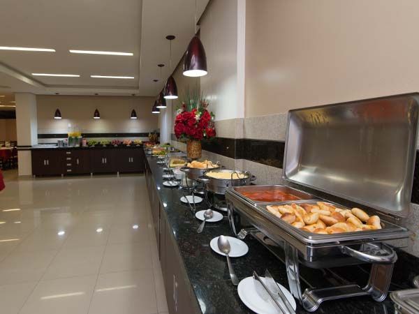 Buffet com comida servida em travessas, iluminação suspensa e balcão comprido, em ambiente interno.