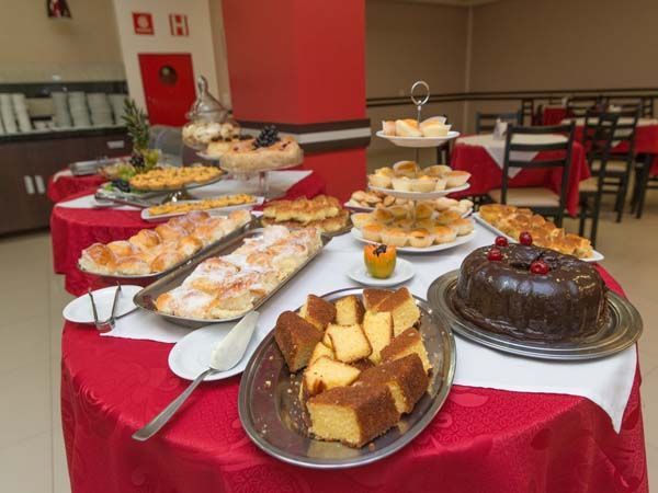 Buffet de sobremesas sobre uma toalha de mesa vermelha. Bolos, doces e outras guloseimas são exibidos em um ambiente de restaurante.