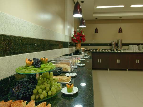 Buffet com diversos itens alimentícios em exposição, incluindo frutas. Balcão escuro, paredes bege e iluminação embutida.