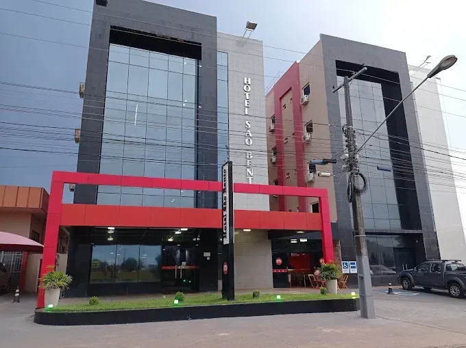 Prédio de hotel moderno com fachada preta, vermelha e de vidro. Iluminação externa, fios elétricos e um caminhão visíveis.