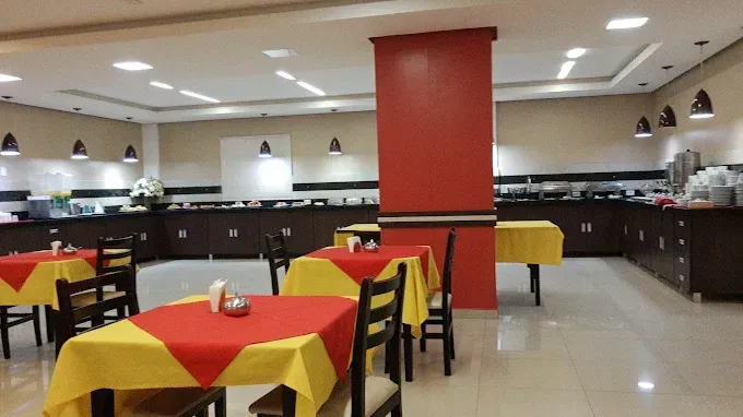 Interior de restaurante com toalhas de mesa amarelas e vermelhas, um bufê e cadeiras pretas.