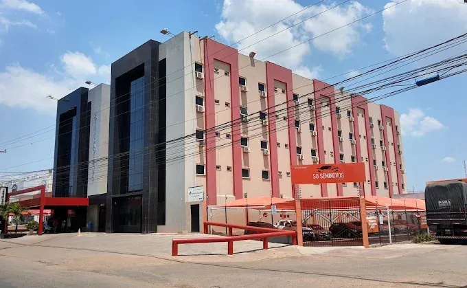 Fachada de hotel com detalhes em vermelho, uma placa e veículos estacionados sob um céu claro.