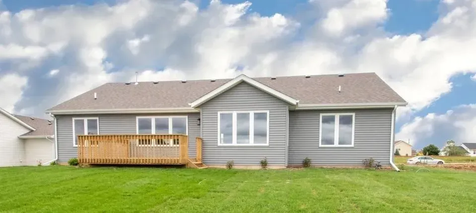 Back of a gray house with a wooden deck, set on a green lawn, under a cloudy blue sky.