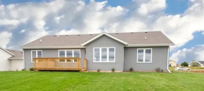 Back of a gray house with a wooden deck and green lawn under a cloudy sky.