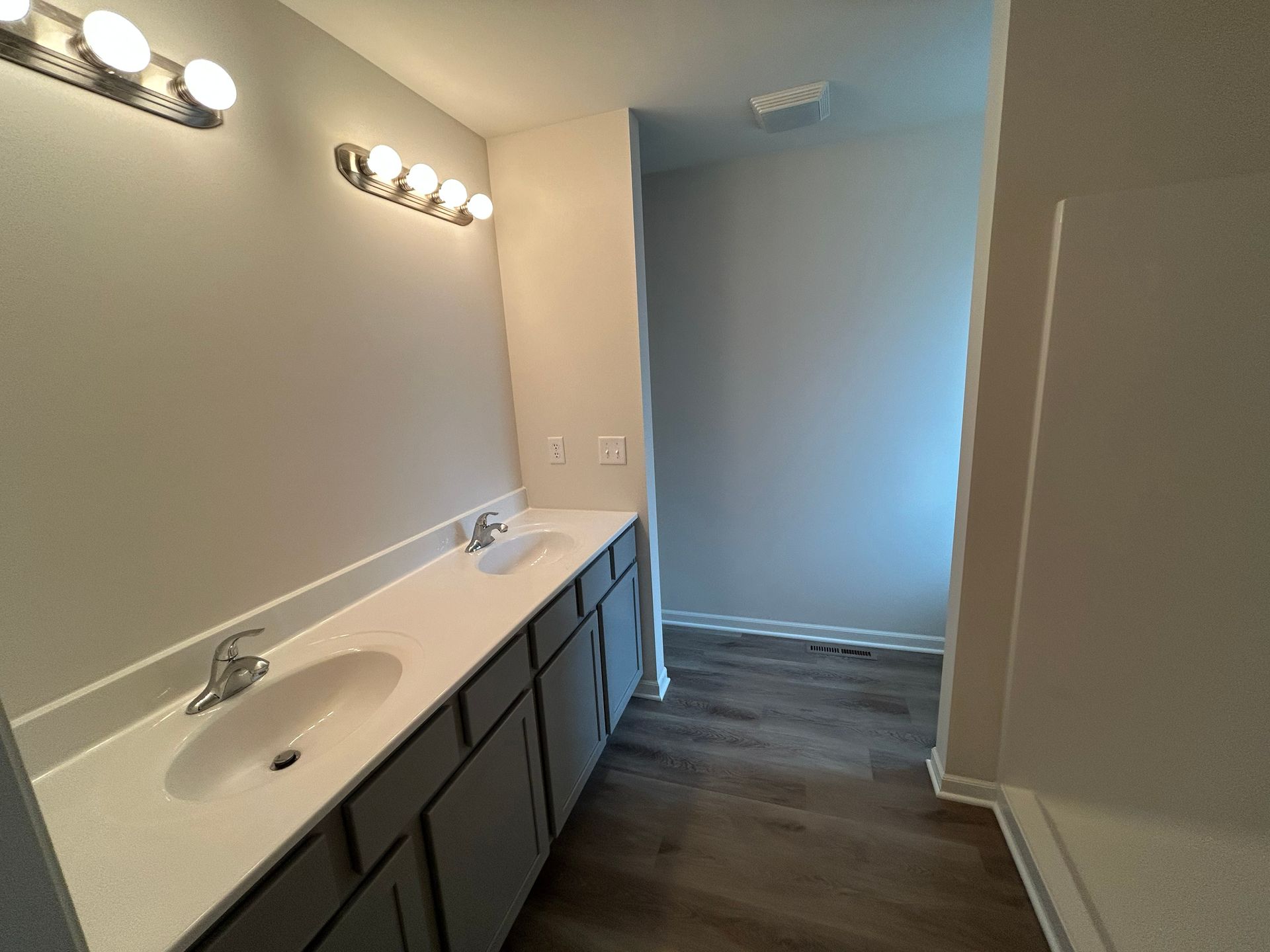Bathroom with gray cabinets, white countertops, dual sinks, and a doorway to the right.