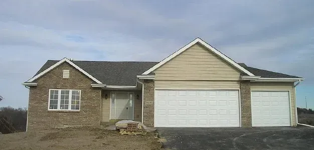 House under construction with brick exterior and two garage doors. Asphalt driveway in front of the house. Cloudy sky.