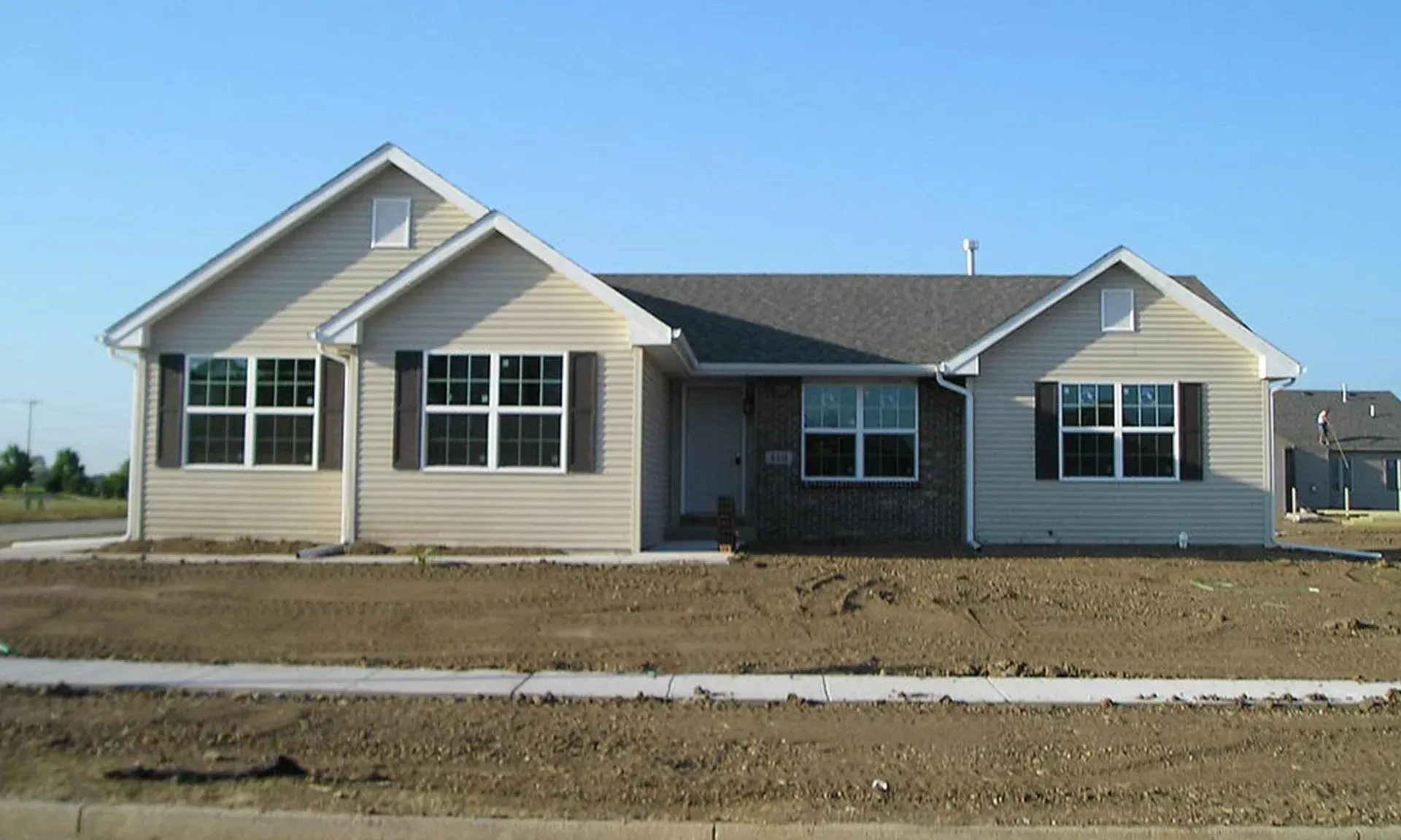 Beige ranch home under a clear blue sky with brown shutters, on a bare lot.