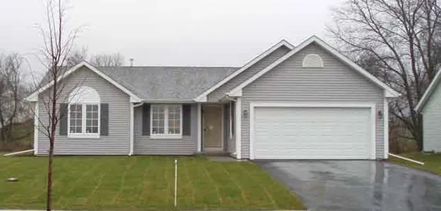 Gray house with a garage, shutters, and a lawn. Overcast sky.