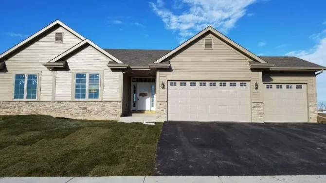 Tan ranch house with two-car garage, stone accents, and dark asphalt driveway on a sunny day.