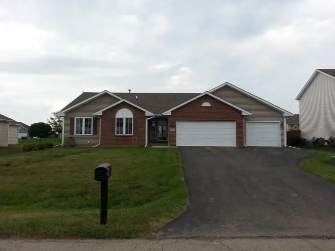 A single-story beige and brick house with a two-car garage on a cloudy day. Black mailbox in front.
