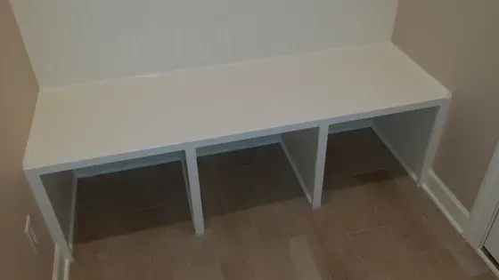 Built-in white bench with three cubby openings in a room with light-colored walls and wood flooring.