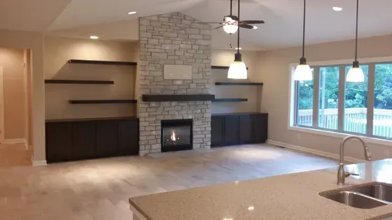 Living room with stone fireplace, built-in shelves, dark cabinets, large windows, and light-colored wood floors.